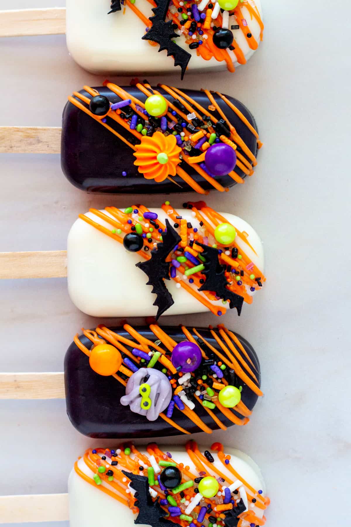 cakesicles lined up, alternating white and black with orange chocolate drizzle and Halloween sprinkles