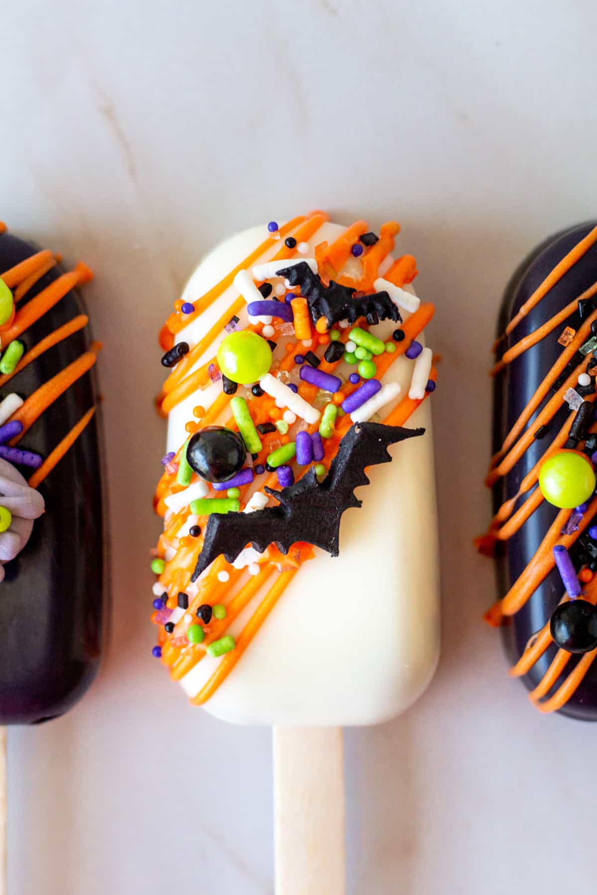 close up of white cakesicle with orange chocolate drizzle and Halloween sprinkles with a fondant bat